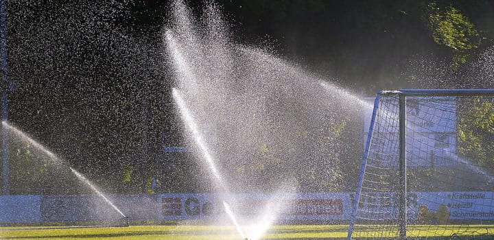 Sprinkler bewässert ein Fußballfeld mit starkem Wasserstrahl neben einem Tor als Motiv für Hochleistungsbewässerung und Sportplatzbewässerung