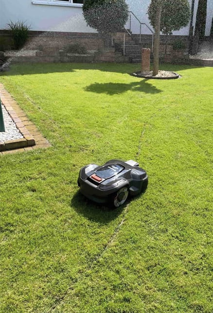 Professionelle Mähroboter auf grünem Rasen während im Hintergrund ein Regner aktiv ist, um das Zusammenspiel von automatischer Bewässerung und autonomer Rasenpflege zu verdeutlichen