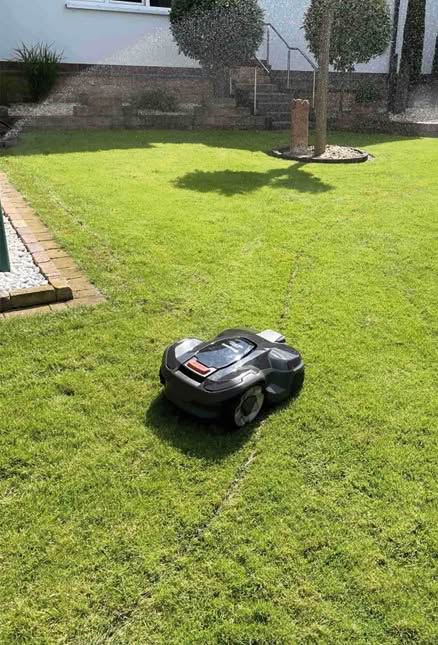 Perfekte Abstimmung zwischen Mähroboter und automatischer Bewässerung Professionelle Mähroboter auf grünem Rasen während im Hintergrund ein Regner aktiv ist, um das Zusammenspiel von automatischer Bewässerung und autonomer Rasenpflege zu verdeutlichen